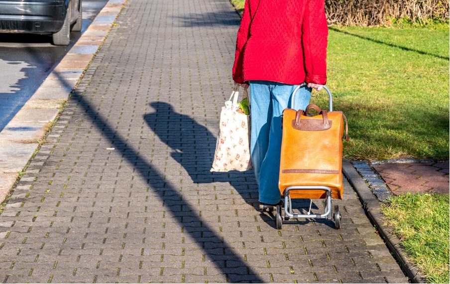 Image of a man or woman walking along a pavement, pulling a trolley and carrying a bag of grocies. The person is wearing a red jacket, and jeans with sandals. 